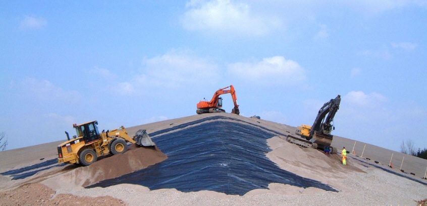 Flera arbetsfordon arbetar med att täcka över en deponi. Täckningsduk syns under lager med sand.