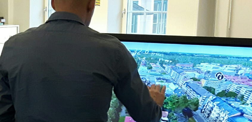 Man står vänd mot digital skärm som visar visualisering av hus och gator i stadsmiljö.