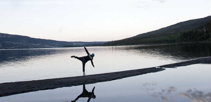 En person balanserar på ett ben, stående på en sten med armarna utsträckta, omgiven av vatten med fjäll i bakgrunden i skymningsljus