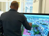 Man står vänd mot digital skärm som visar visualisering av hus och gator i stadsmiljö.
