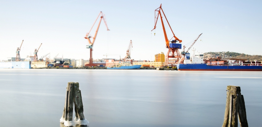 I förgrunden två pålar som sticker upp ur vattnet och i bakgrunden Göteborgs hamn med skepp och lyftkranar. 