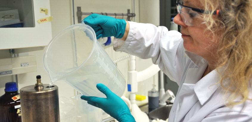 Kvinna i laboratorierock och handskar häller vatten från en tillbringare ner i en behållare som tillhör laktestförsöket.